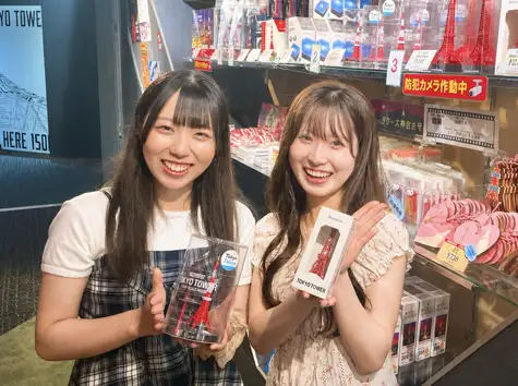 東京タワーのミニチュア模型を手に持って笑顔を見せる2人の女性と、その背後に並ぶお守りやグッズの数々