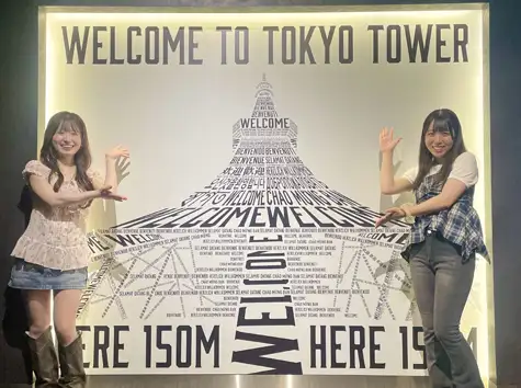 「WELCOME TO TOKYO TOWER」の文字と世界各国の「ようこそ」の言葉でタワー型に飾られた東京タワーメインデッキのフォトスポットで、笑顔で写真を撮り合う2人の女性